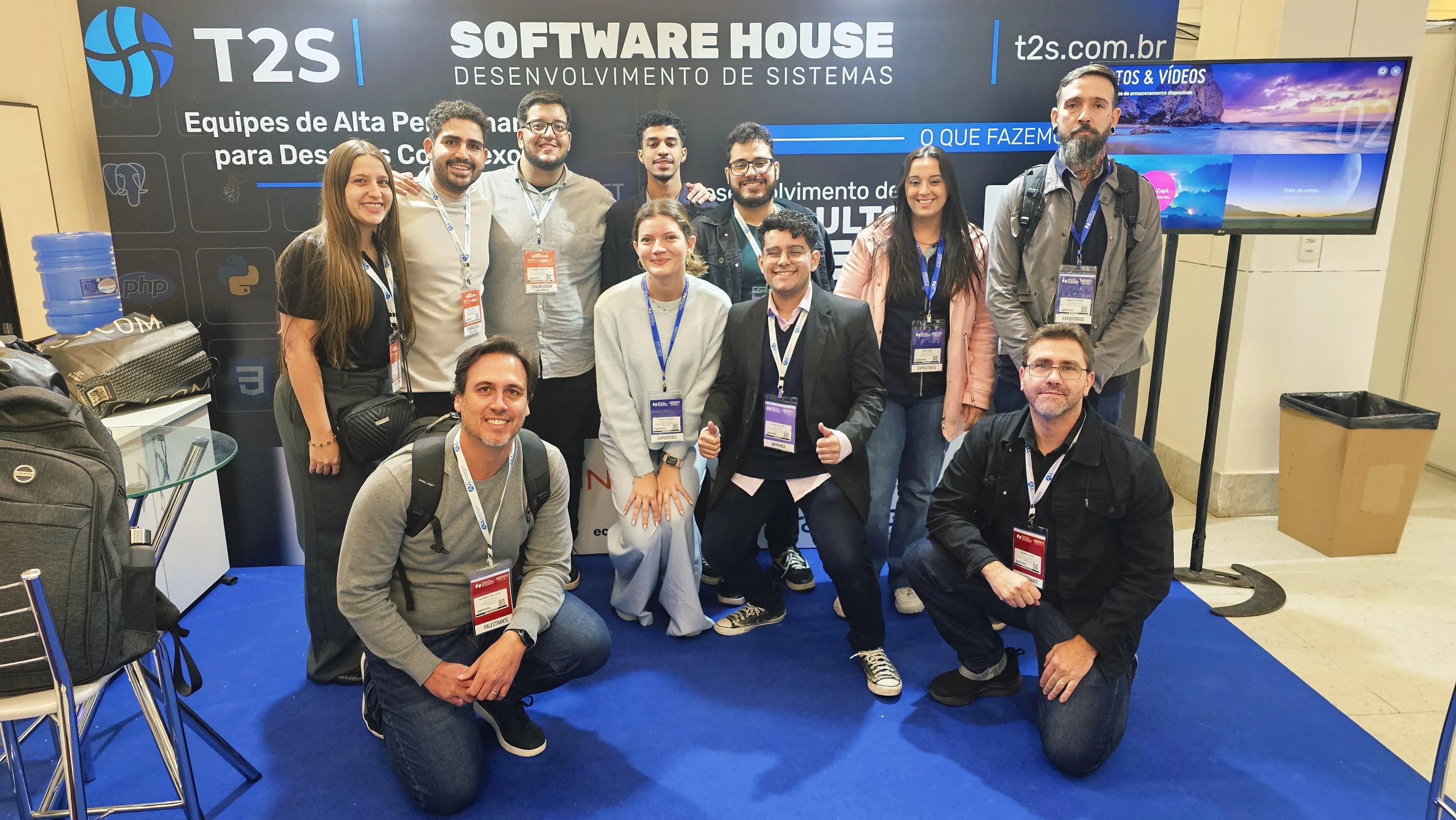 Equipe da T2S Software House reunida em estande de evento, com banner destacando tecnologias como React, Python e PHP ao fundo.